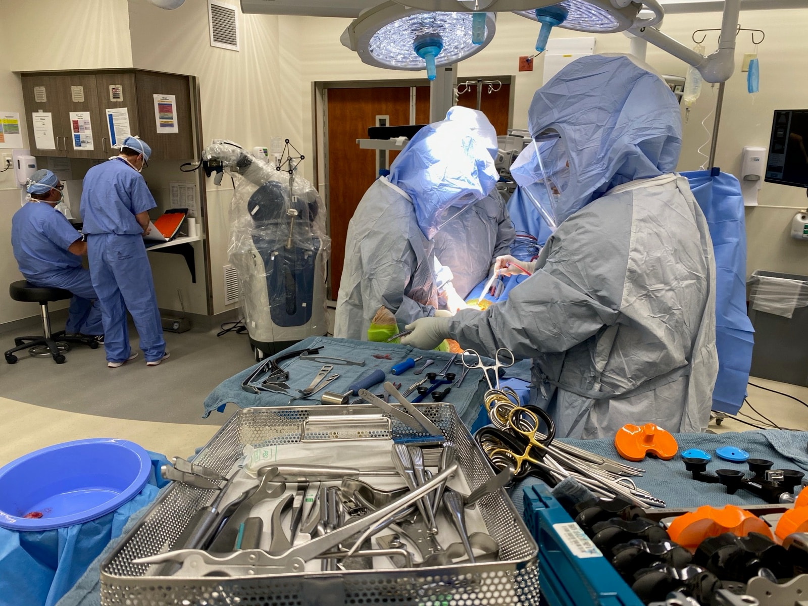 Dr. Jason Jones and a colleague in surgical helmets operating on a knee, the draped Mako robotic arm behind them and instrument trays in front