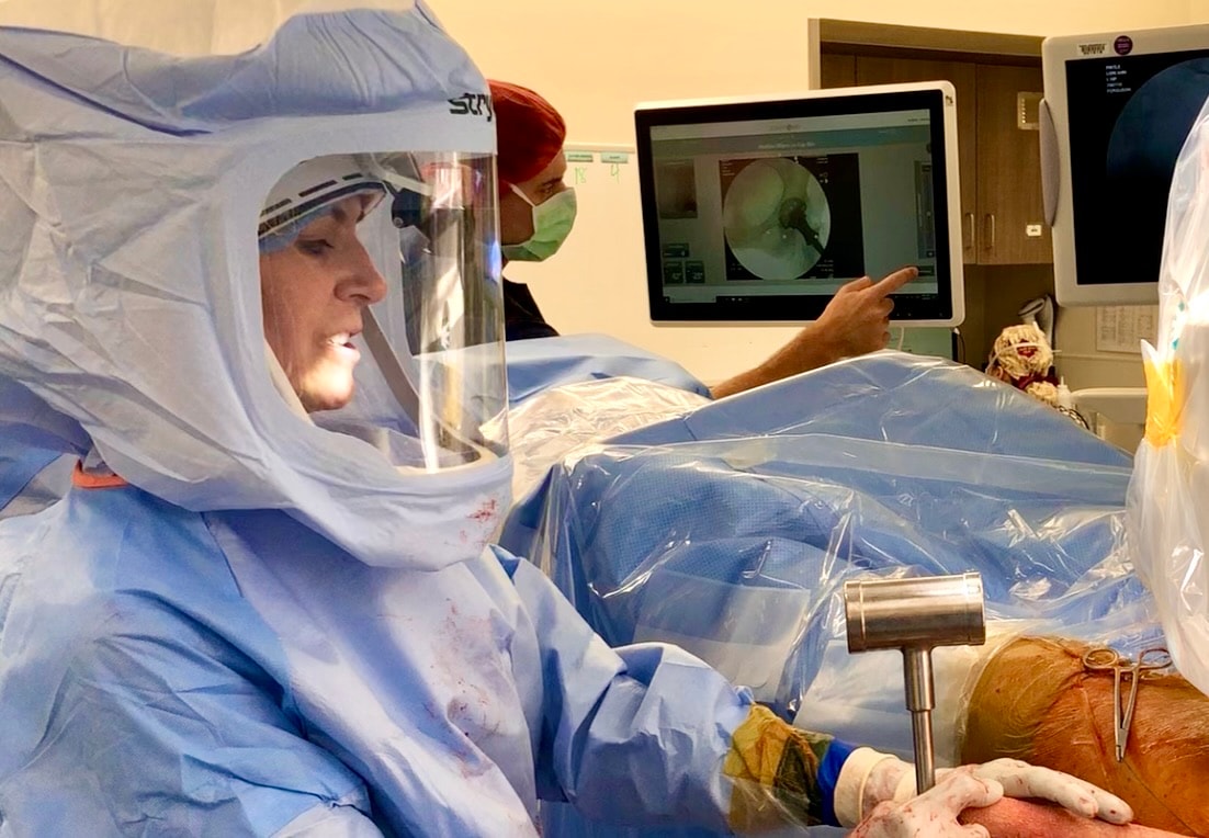 Dr. Tania Ferguson in a surgical helmet during hip surgery, as a colleague points to the live X-ray on the monitor