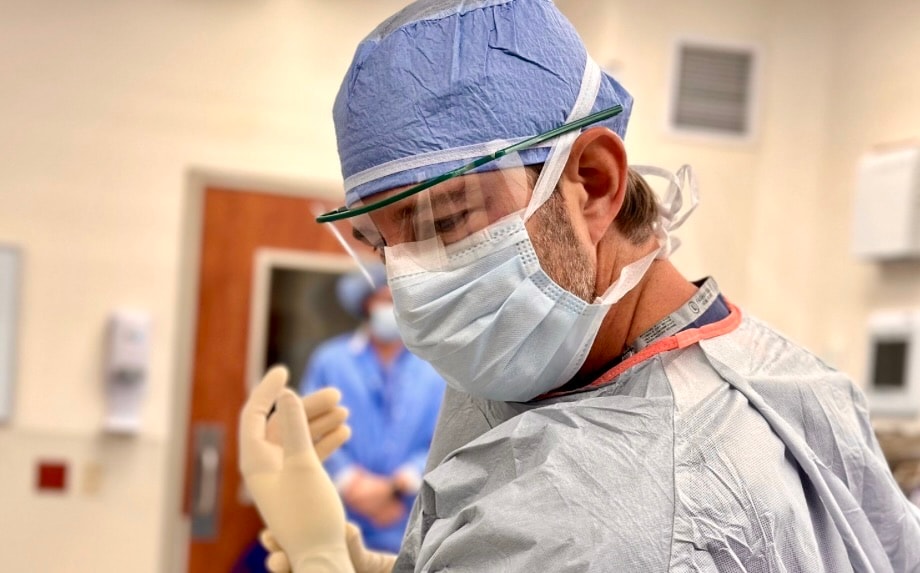 Dr. Jason Jones in cap, mask and eye shield, pulling on surgical gloves before an operation